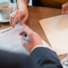 Close up man hand signing contract with female partner. They tasting cup of coffee while situating at desk. Business concept